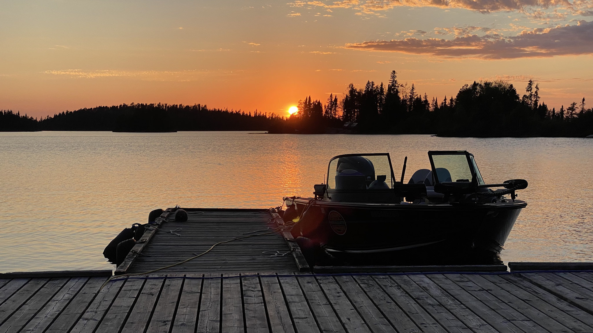 a motorboat next to a wooden dock, silhouetted by an orange sunset over a calm lake.
