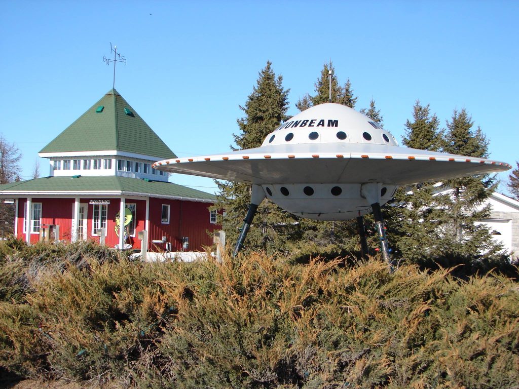 a large sculpture of a flying saucer with the word "Moonbeam" on the side, raised on stilts so that it appears to float above the trees. It stands next to an ornate red and green heritage building under a clear blue sky.