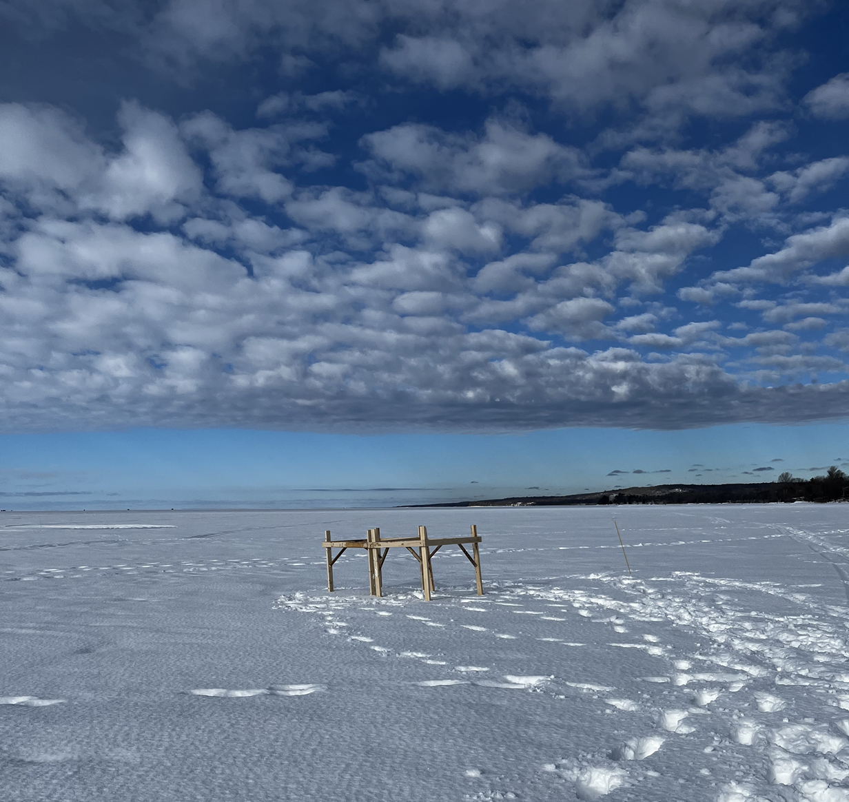 Une base de bois repose sur le lac gelé et enneigé. Des nuages en forme de boules de coton parsèment un ciel bleu clair.