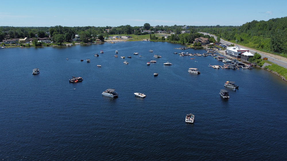 An overhead shot of the wide blue Trout Lake, dotted with many boats that are there for the Boatapalooza waterfront concert. On the shore in the distance there are docks and Average Joe's, the restaurant/ music venue. The sun is shining and the sky is clear blue.