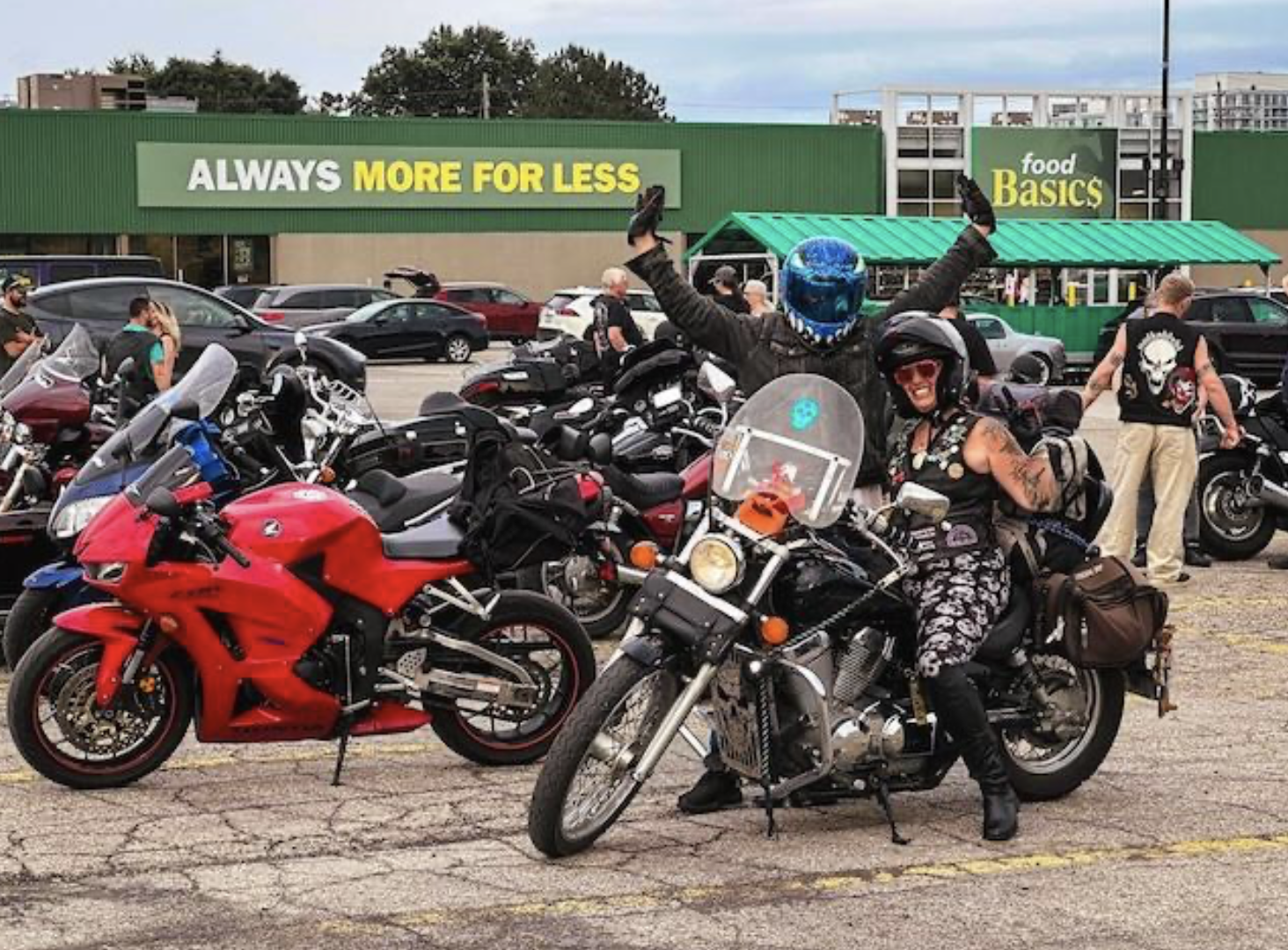 Two motorcyclists smiling and flexing on their motorbikes in a grocery store parking lot full of motorcycles.