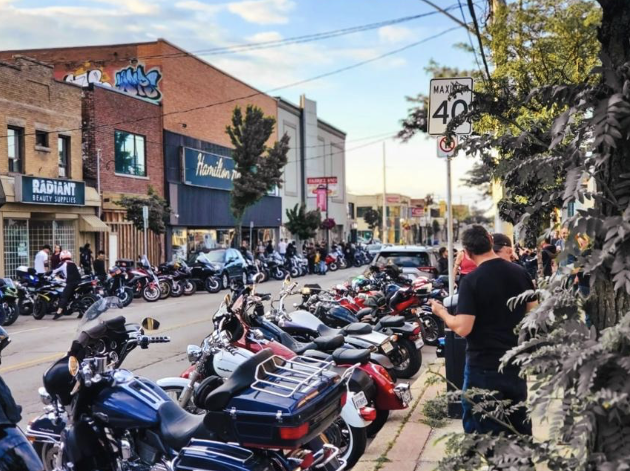 A quiet Hamilton street lined on both sides with parked motorcycles on a summer evening. People are chatting on the sidewalk next to their bikes.