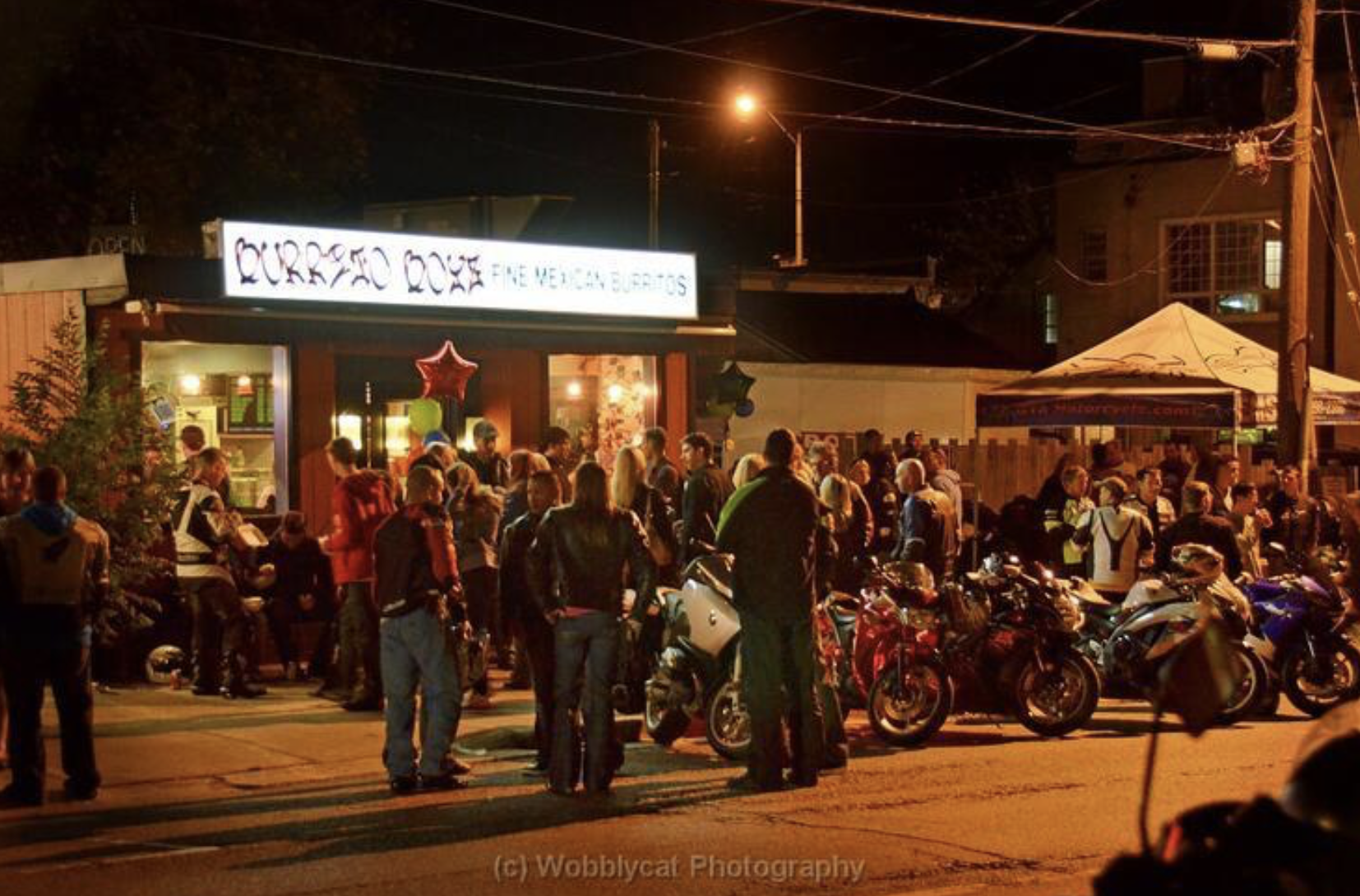 A crowd of people with their motorbikes hanging out and talking in front of an illuminated Burrito Boys restaurant at night.