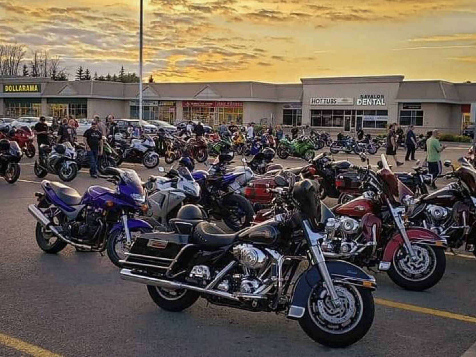 A strip mall parking lot filled with rows of parked motorcycles and groups of people standing around visiting under a golden sunset on a summer evening.