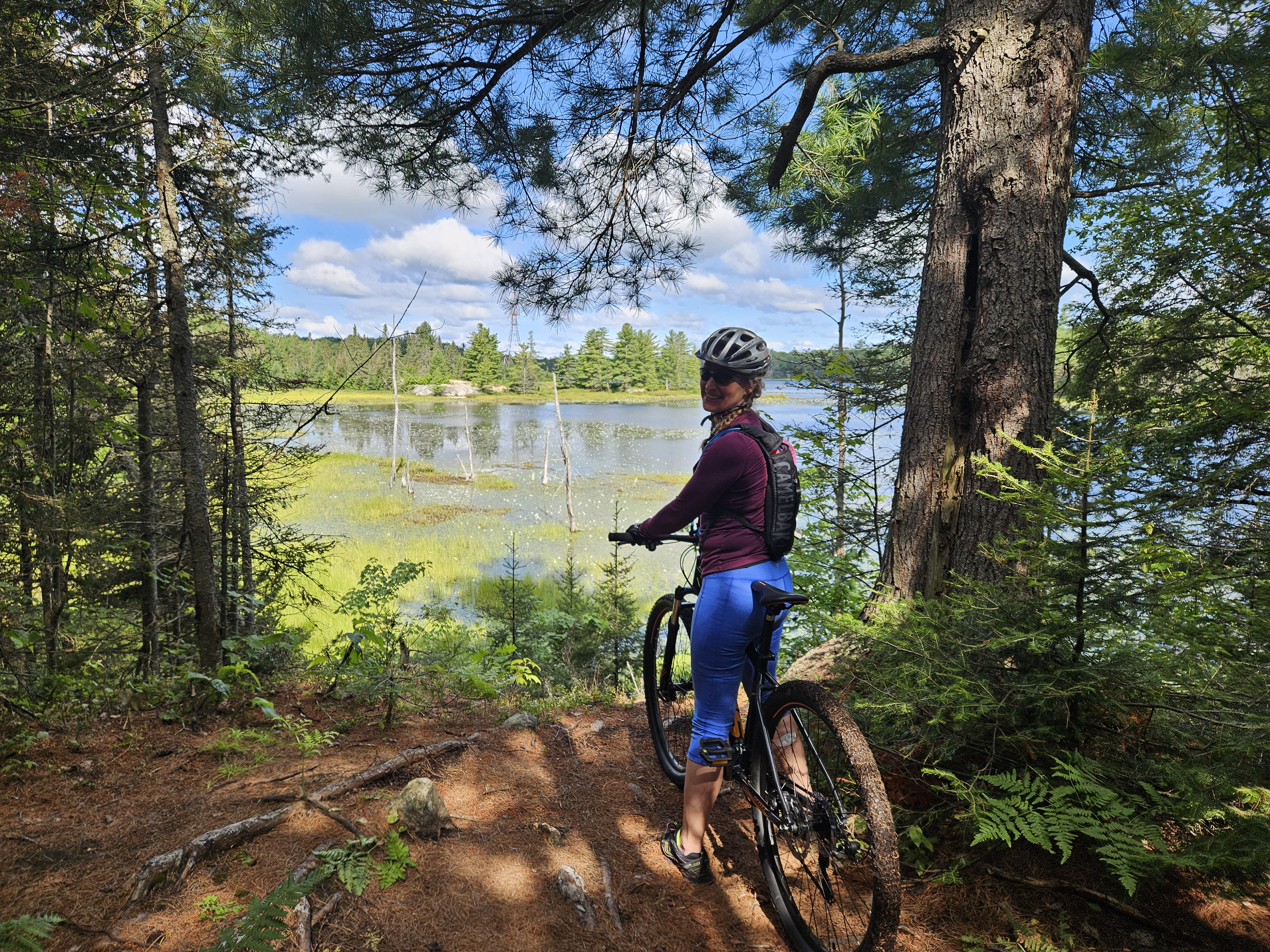A cyclist stands in front of a shining lake in a clearing along a forested biking trail on a sunny day.
