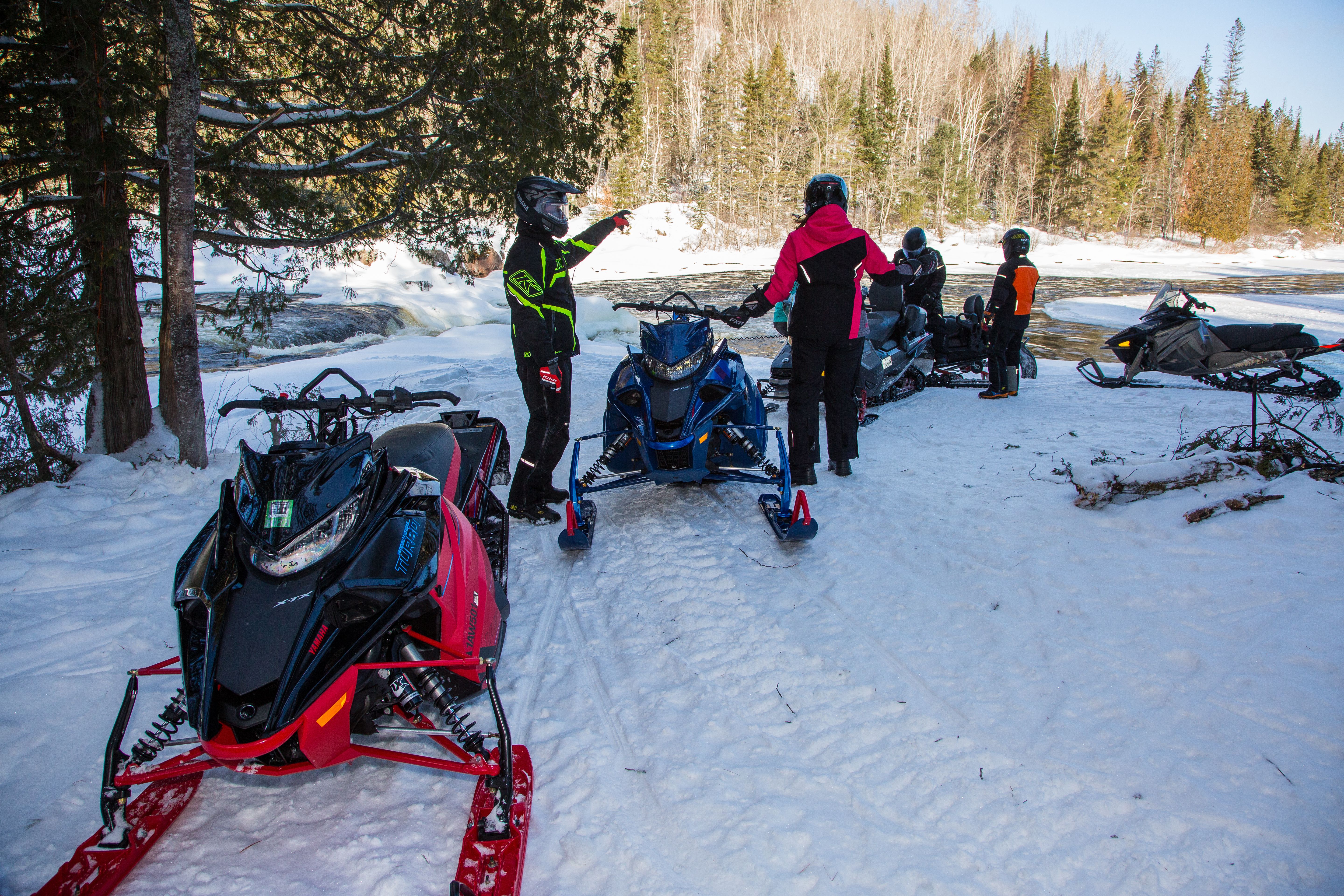 A group of snowmobilers chat next to their machines on a snowy trail on a sunny winter day.