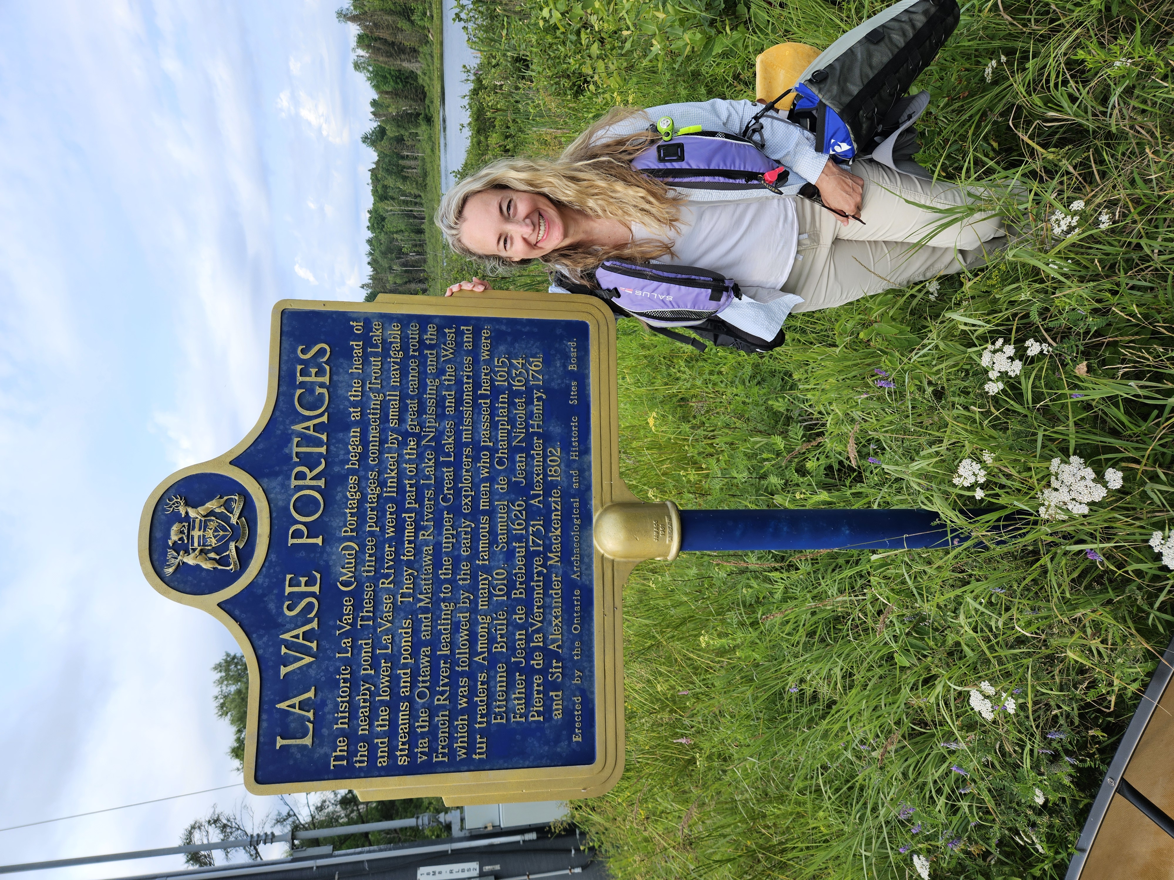 A woman standing in waste-high grass next to an informational sign about the La Vase Portages.