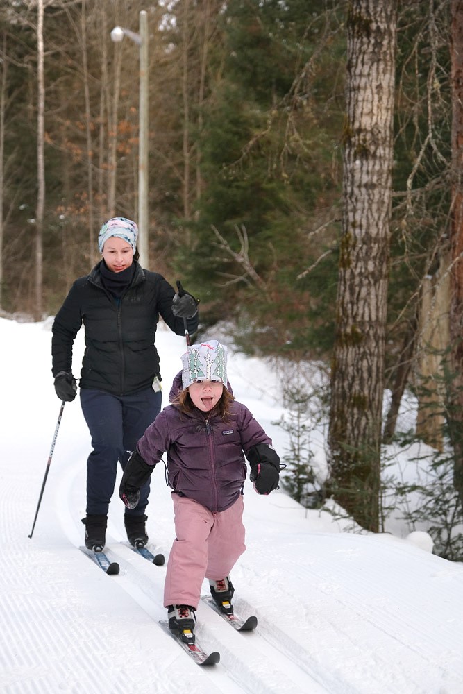 Une petite fille, suivie par une adulte, est toute souriante en faisant du ski de fond. 