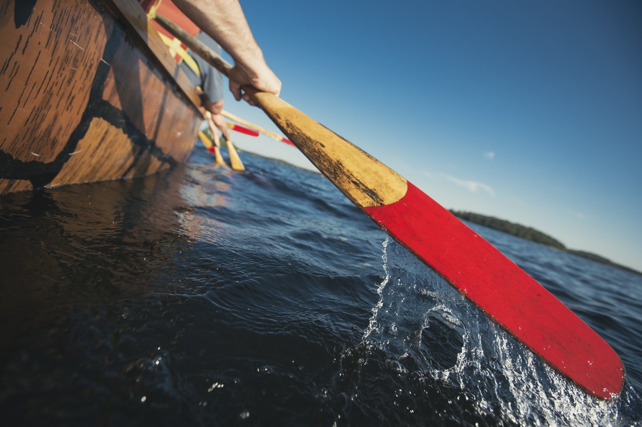 Gros plan sur une pagaie, qui sort de l'eau. Des gens avancent sur l'eau à bord d'un canot voyageur.