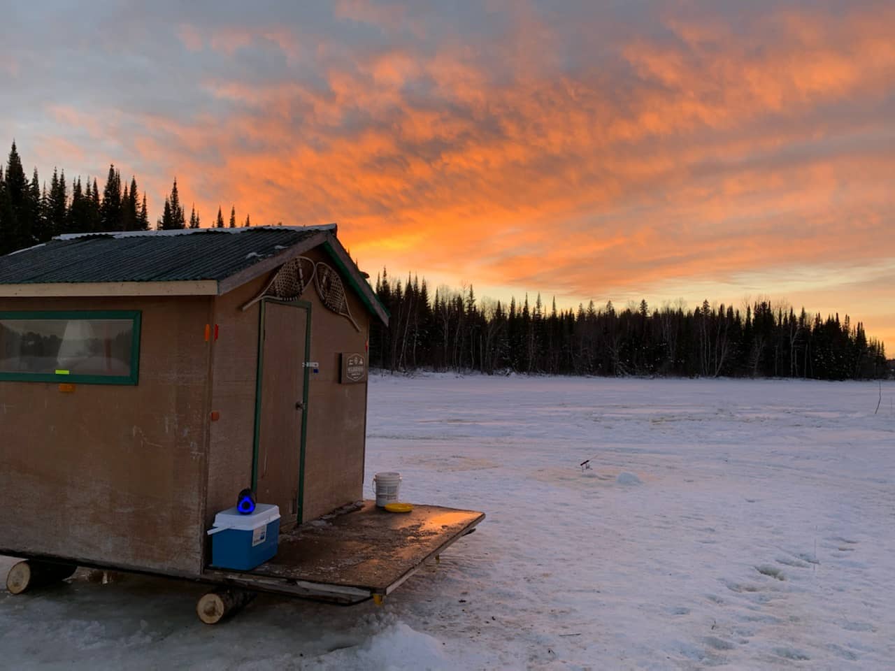 Une cabane de pêche est installée sur un lac. Le soleil se couche : les nuages sont orangés.