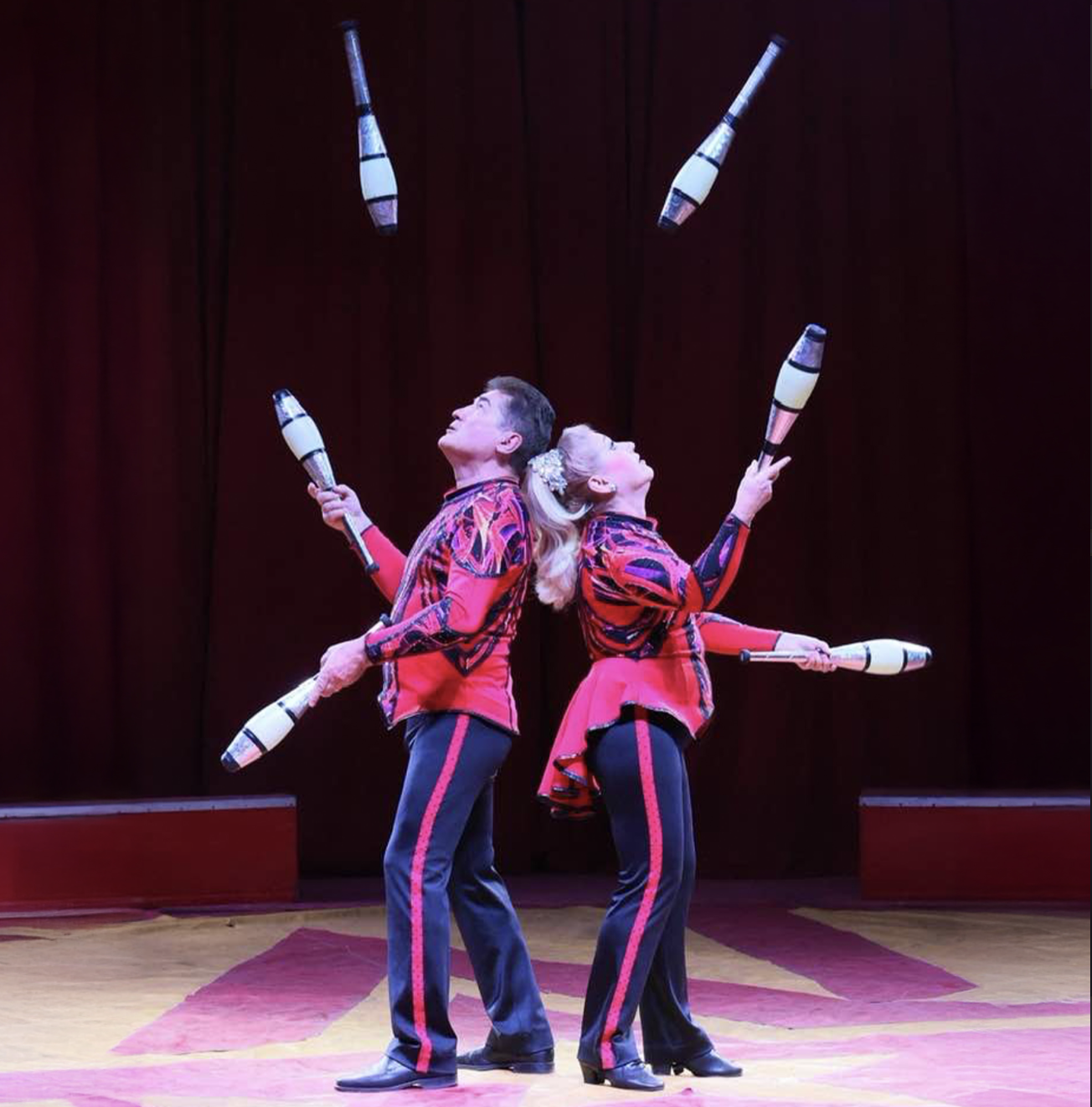 two jugglers stand back to back, tossing bowling pins in a high arch over their heads to one another.
