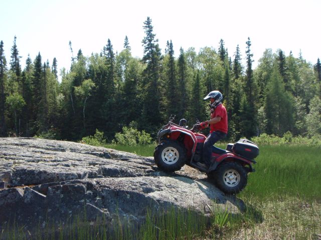 A rider on an ATV drives up a smooth rock hill in the middle of a green grassy clearing in the boreal forest on a sunny summer day. 