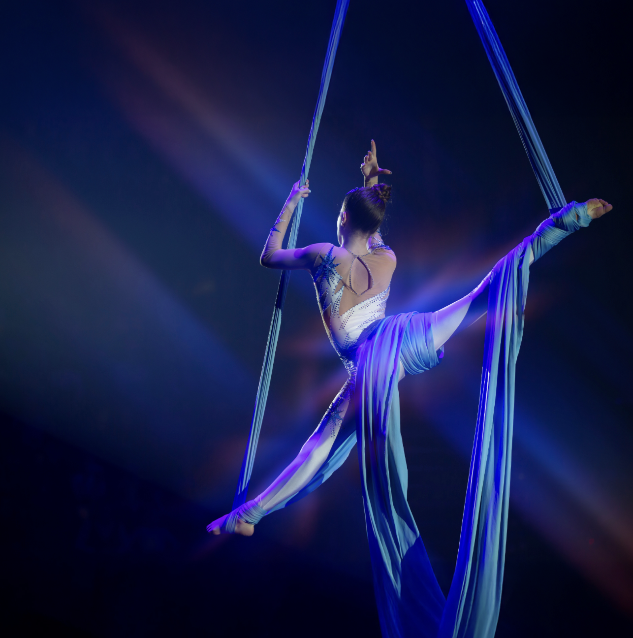 A woman supspended gracefully in a full split position, hanging high above the crowd under blue spotlights by gymnastics silks wrapped and draped around her legs.