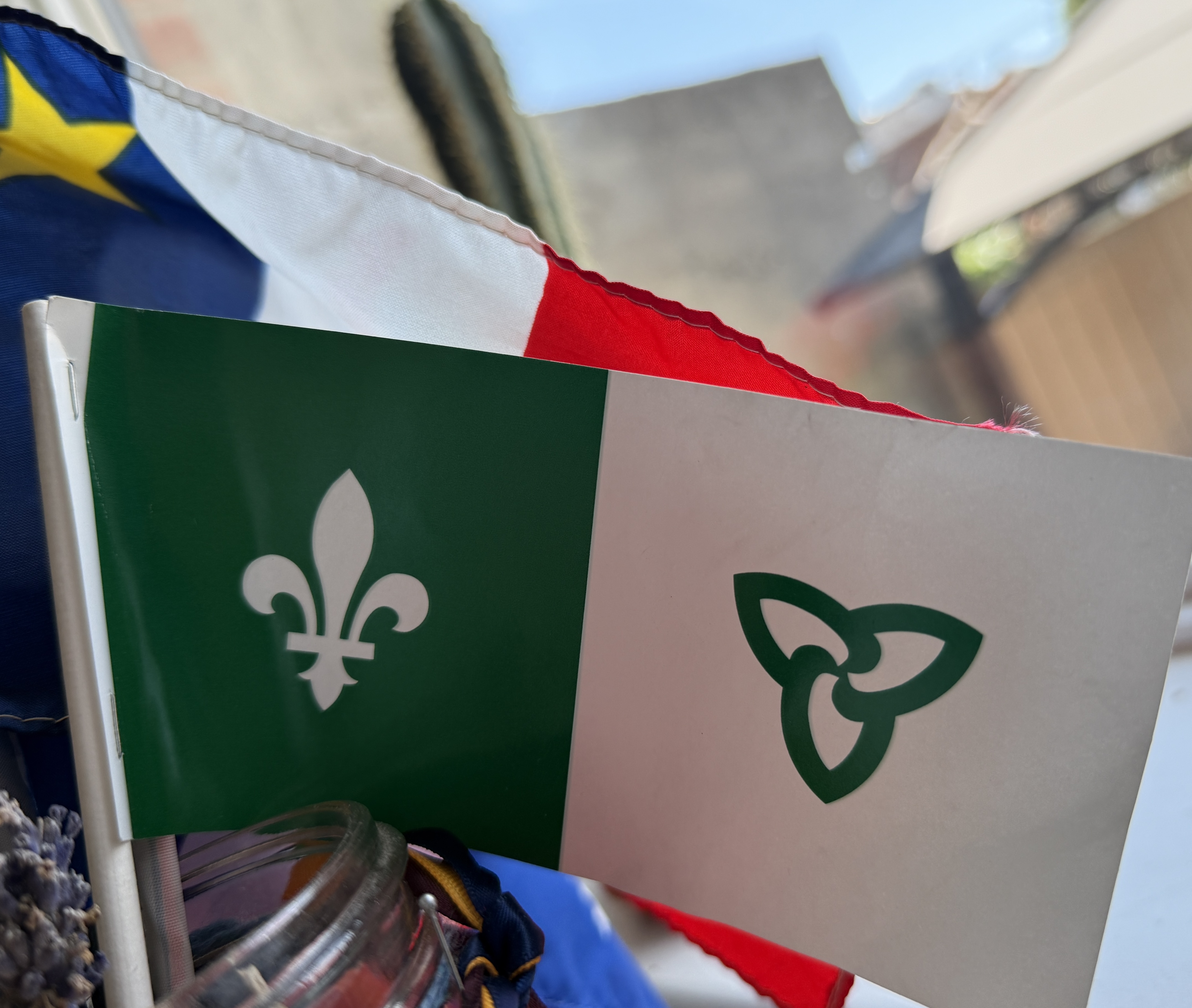 un petit drapeau franco-ontarien de la taille d'une main à côté d'un petit drapeau acadien, tenus ensemble devant un arrière-plan de ciel bleu et de ville.