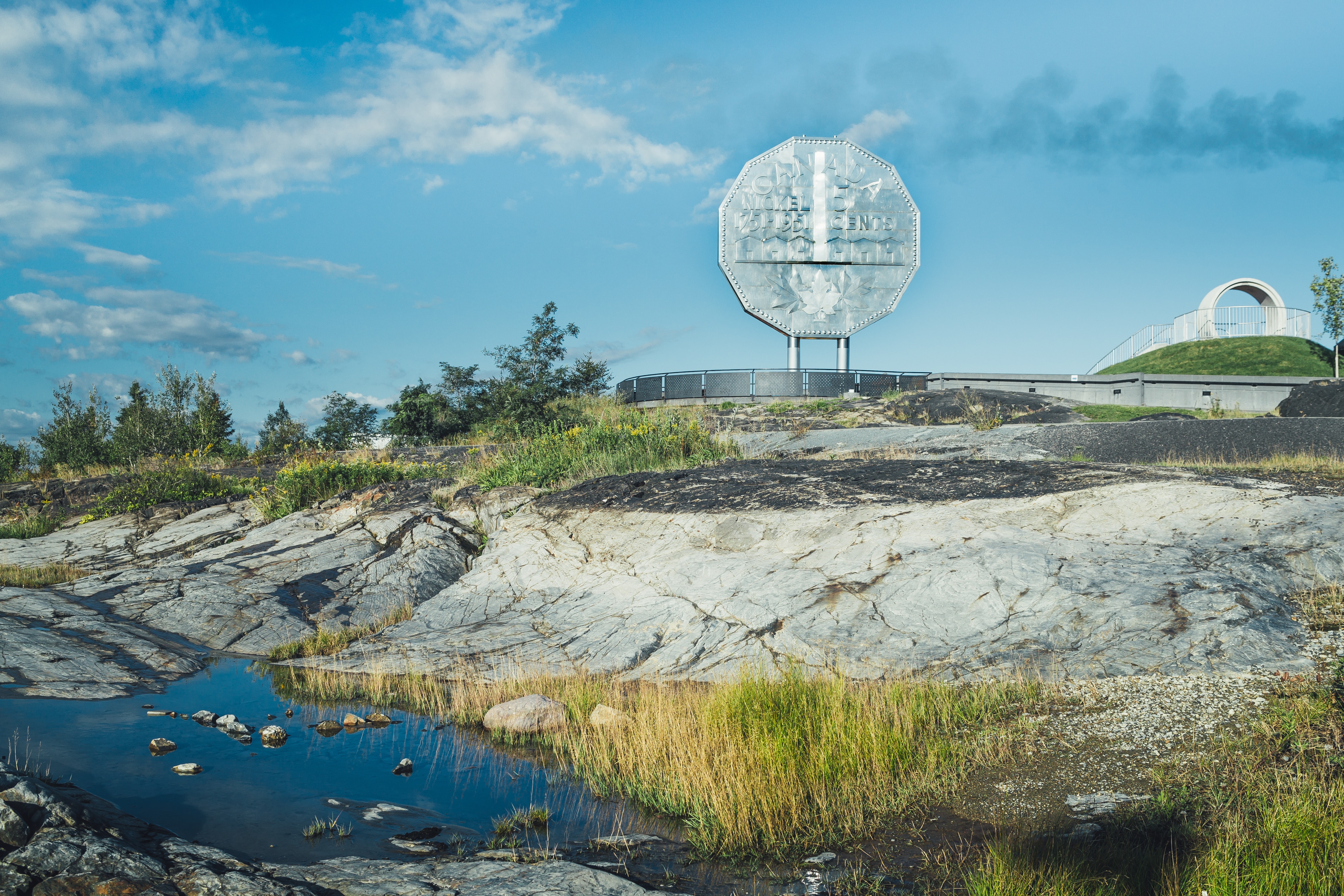 La statue Big Nickel de Sudbury se dresse, brillante, sur la crête d'une colline devant un ciel bleu d'été, avec des bancs de roches lisses et des herbes vertes tout autour.