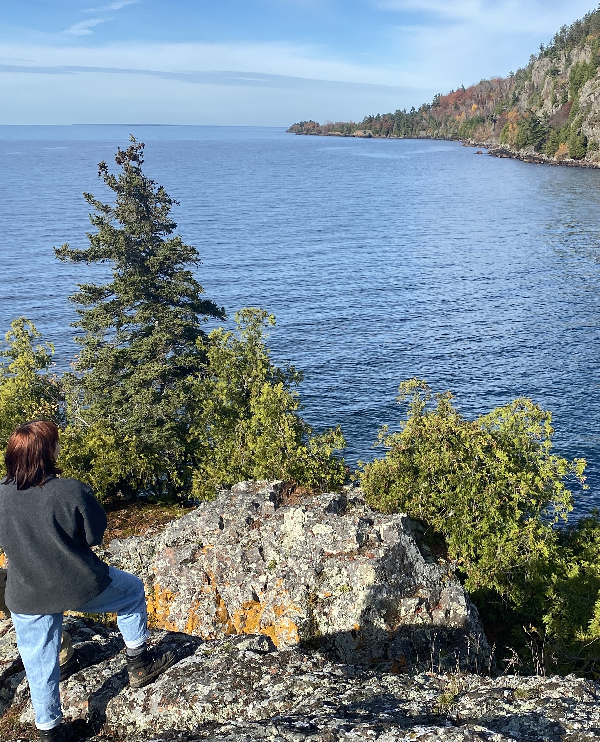 une femme se tient sur un rocher d'observation regardant un lac bleu et une forêt s'étendant devant elle à Gros Cap Bluffs.