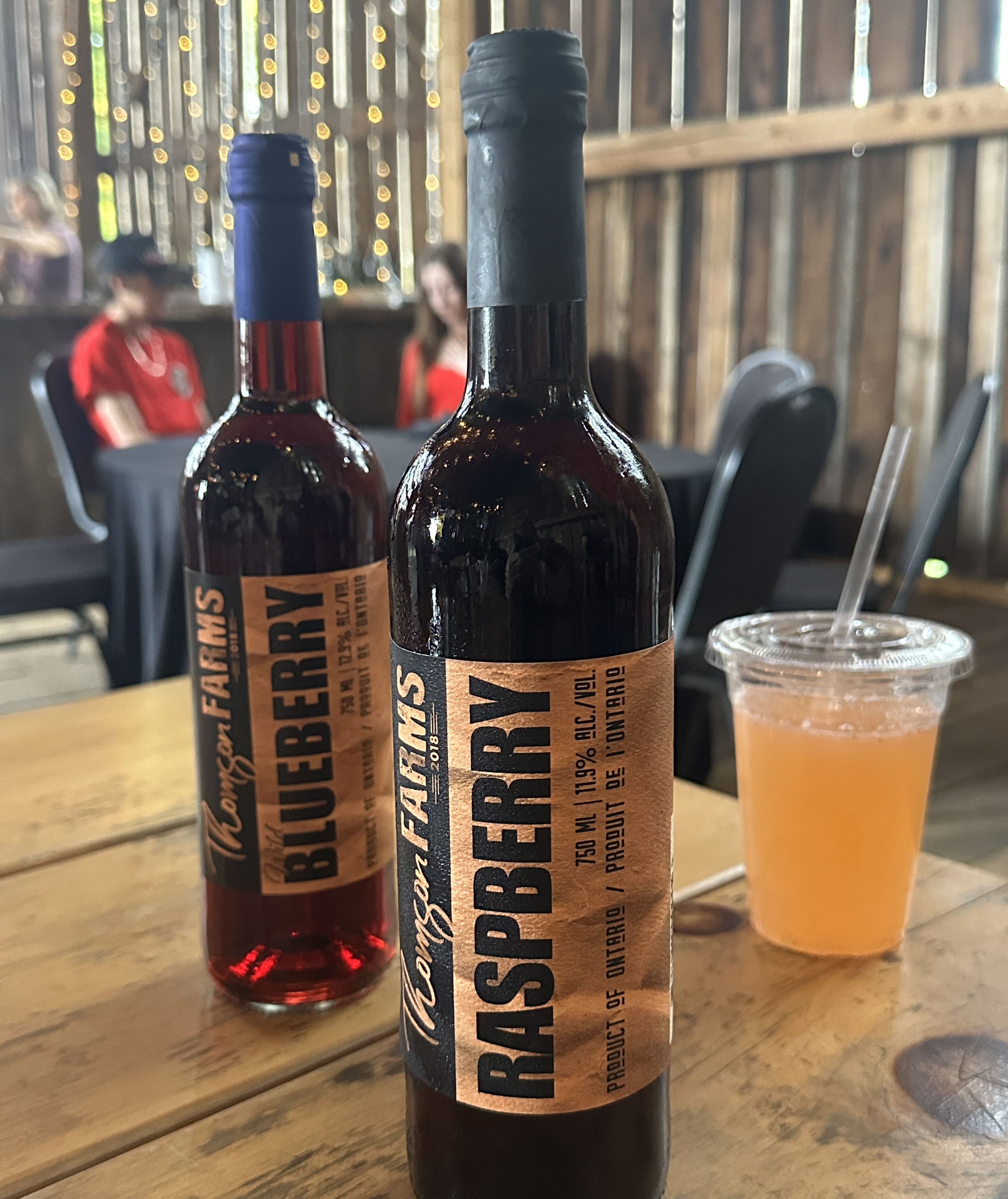 des bouteilles de vin de fruits fabriquées à Thompson Farms sont posées sur une table tandis que des visiteurs sont assis à des tables en arrière-plan.