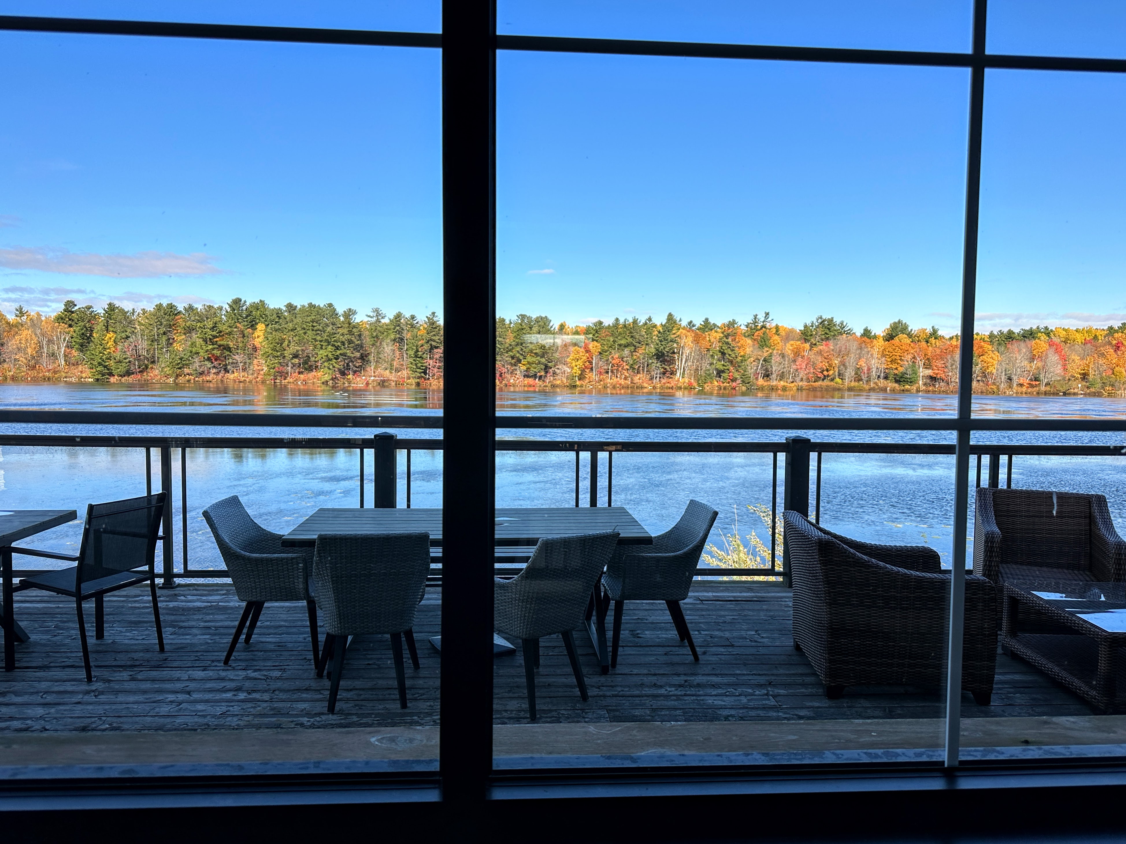 une terrasse extérieure avec de nombreuses tables et chaises, le long d'une rivière calme et scintillante, sous un ciel d'automne d'un bleu éclatant.