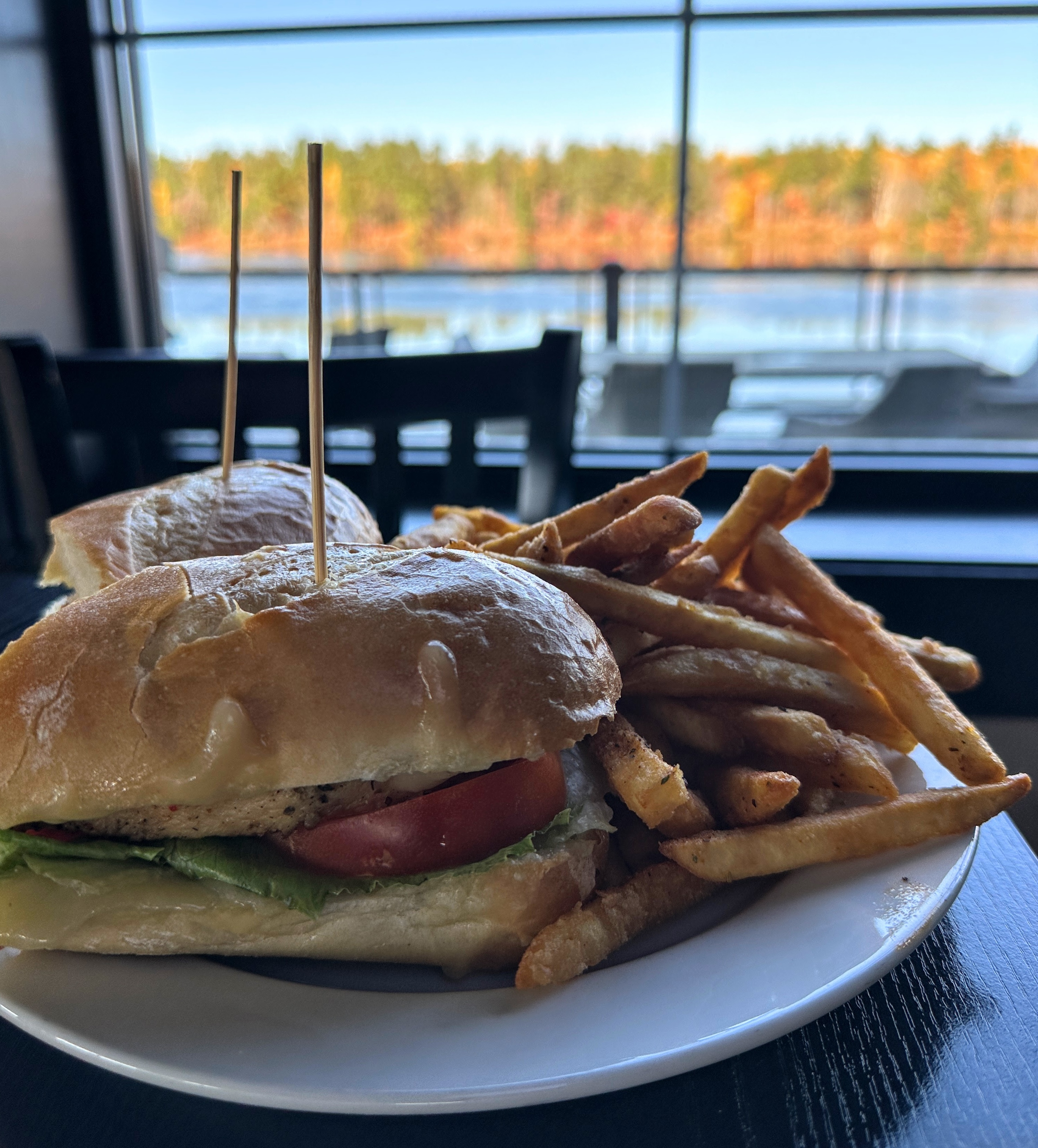 Un sandwich et des frites sont posés sur une table près d'une fenêtre donnant sur une rivière scintillante et des arbres d'automne.