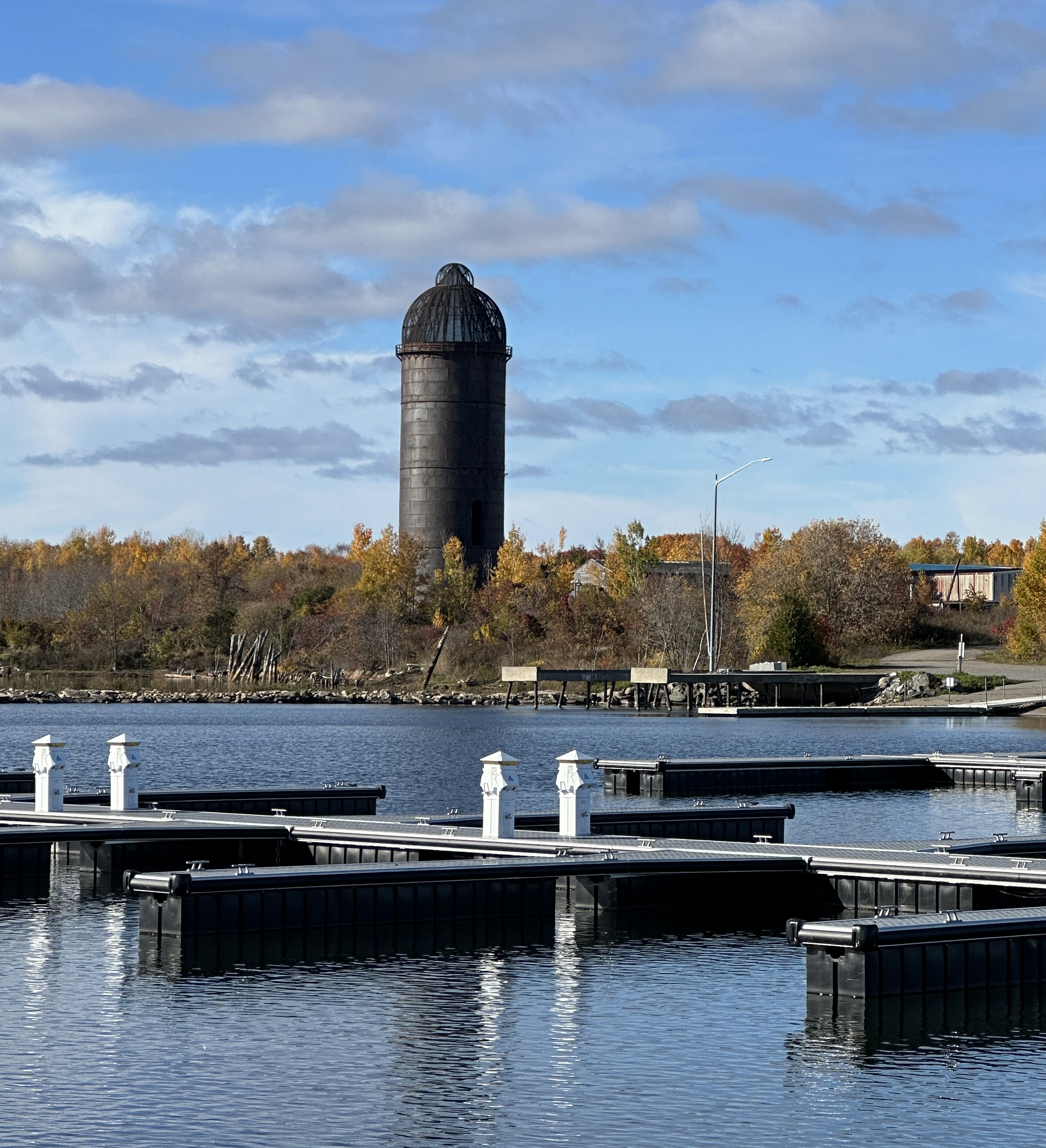 Une imposante tour en métal sombre se dresse au-dessus d'une forêt d'automne aux magnifiques couleurs, au bord d'une rivière scintillante et d'un port de plaisance.
