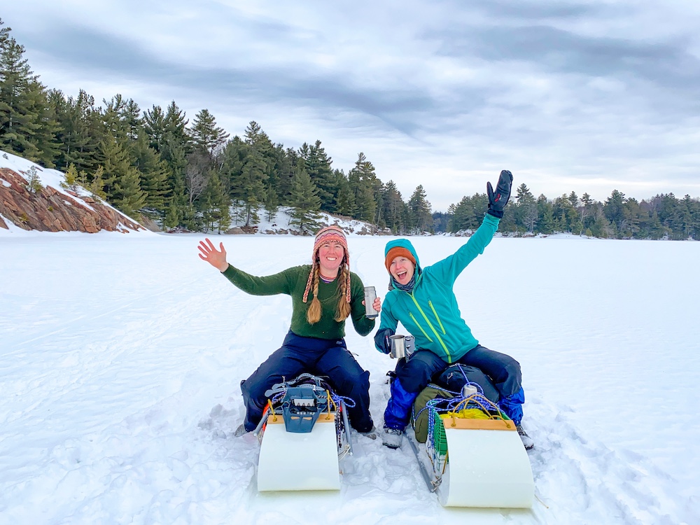 Two women sitting on sleds with winter camping gear on them on a lake in the winter time.