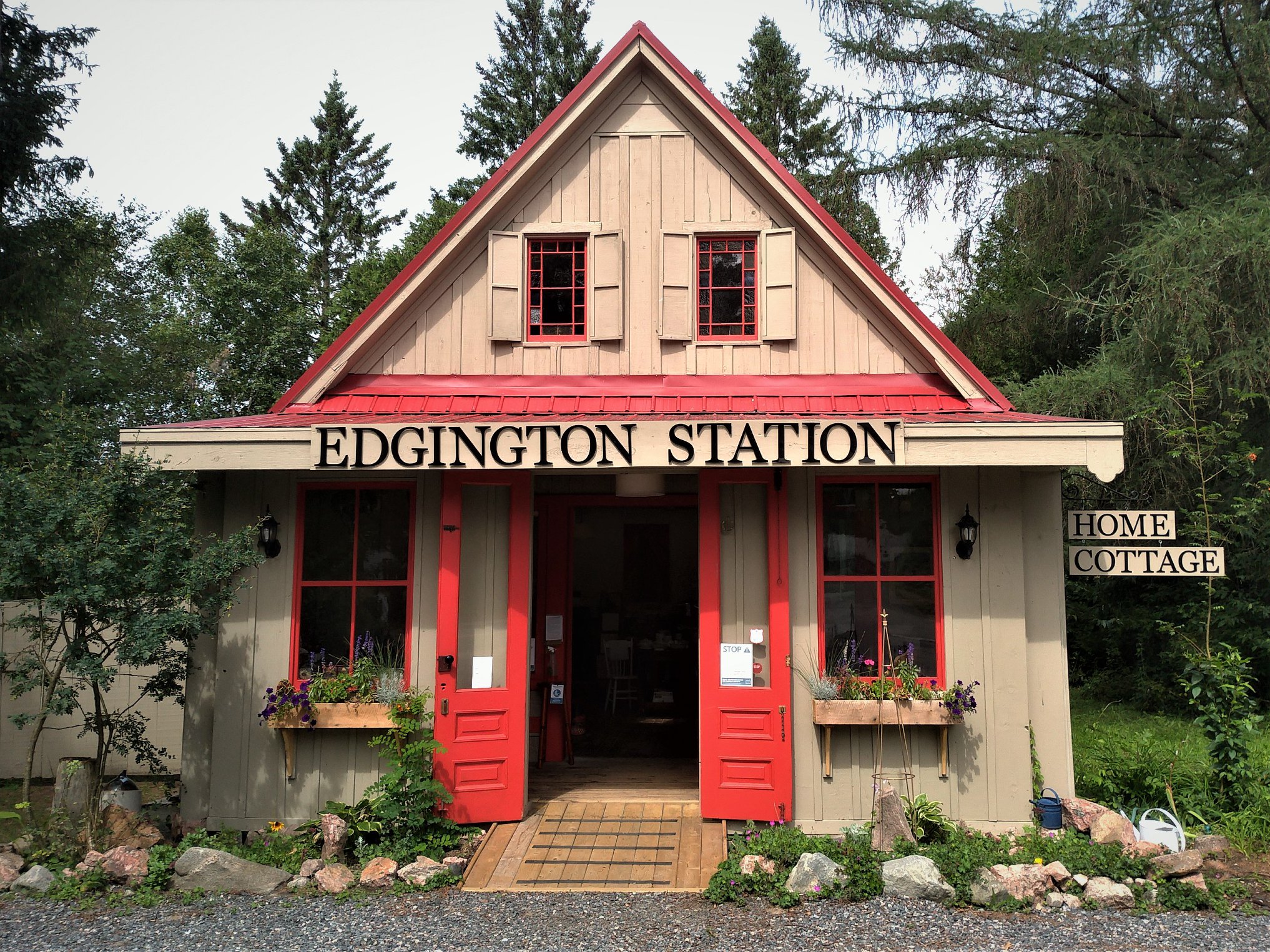 a very cute, trim little old-fashioned shop building with light brown walls and red doors and gables, nestled in some large, aged green trees. The sign above the door reads "Edgington Station".