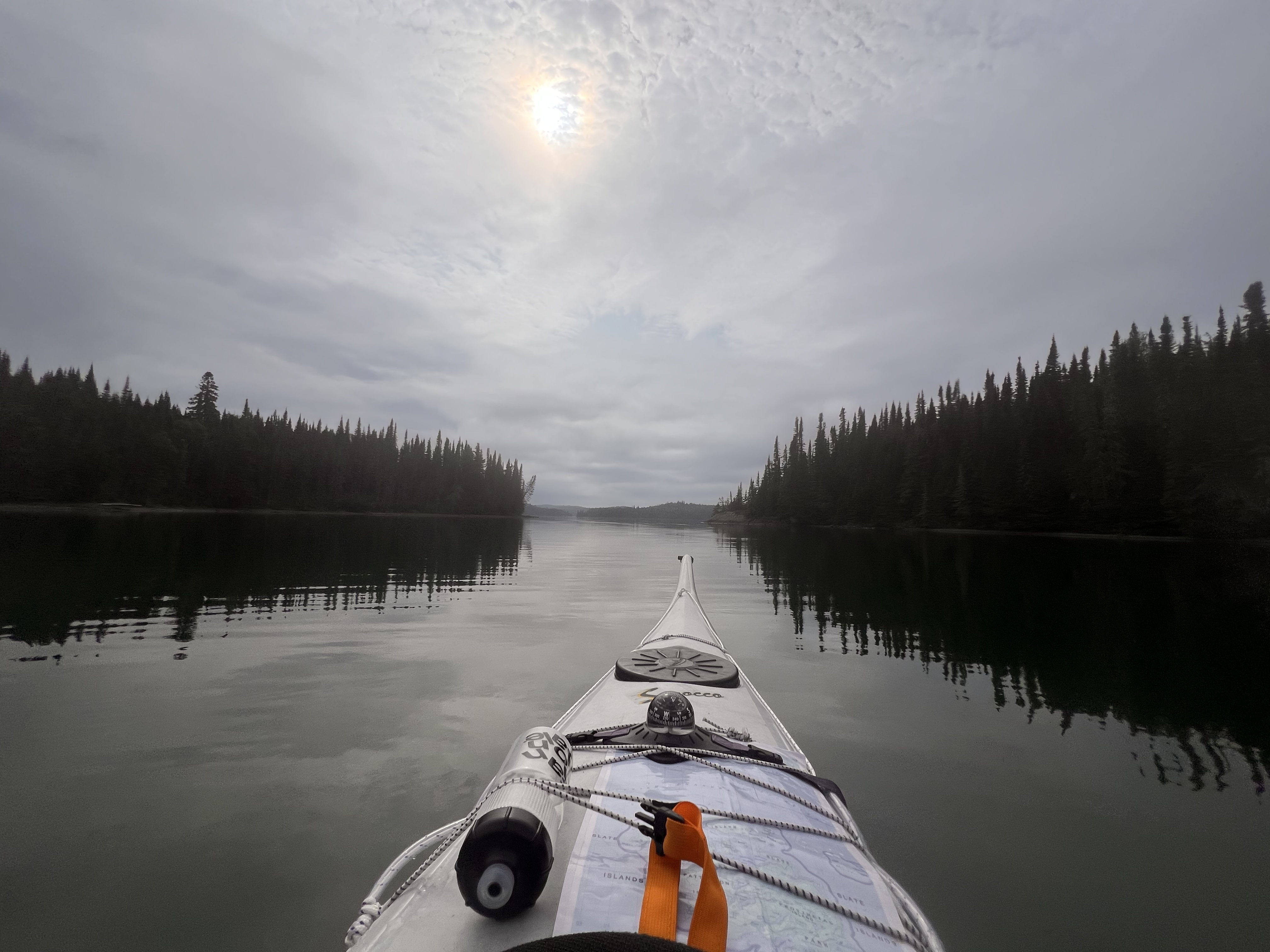 Un kayak, avec une bouteille d'eau et une carte attachées à l'avant, glisse sur les eaux grises et lisses du lac, entre les languettes boisées des îles d'ardoise.