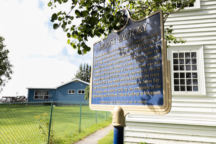 Un panneau d'interprétation sur l'histoire de Moose Factory, devant des bâtiments patrimoniaux en bois.