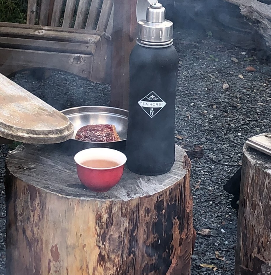 Une petite tasse de thé fumant repose sur une souche près d'un feu de camp, à côté d'un thermos noir étiqueté « Tea Horse ».