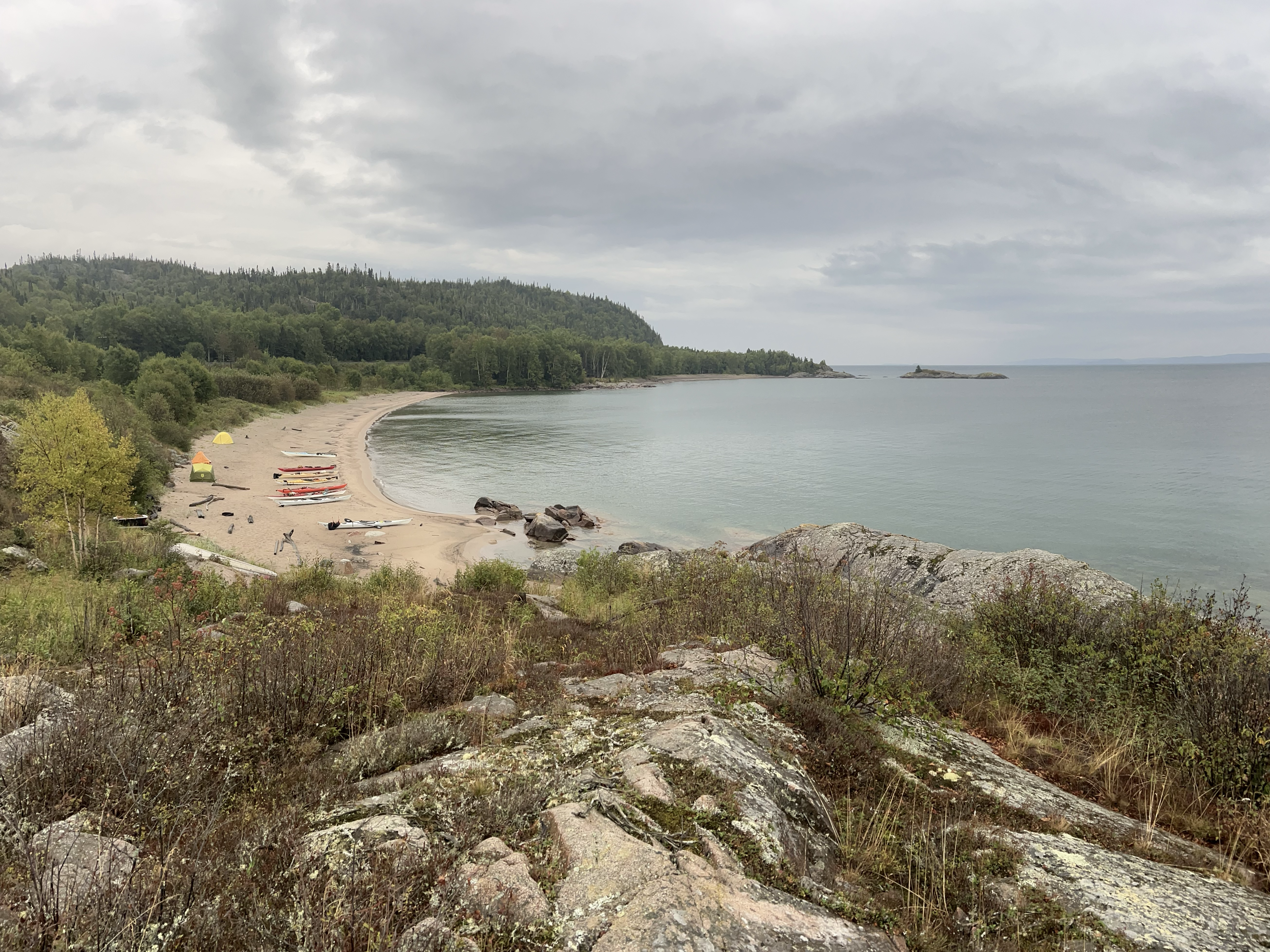 Un groupe de kayaks et de tentes sur une plage de sable de la baie de Jackfish, sur le lac Supérieur. La côte escarpée borde les eaux bleu-gris, tandis qu'une voie ferrée serpente à travers l'épaisse forêt verdoyante au loin.