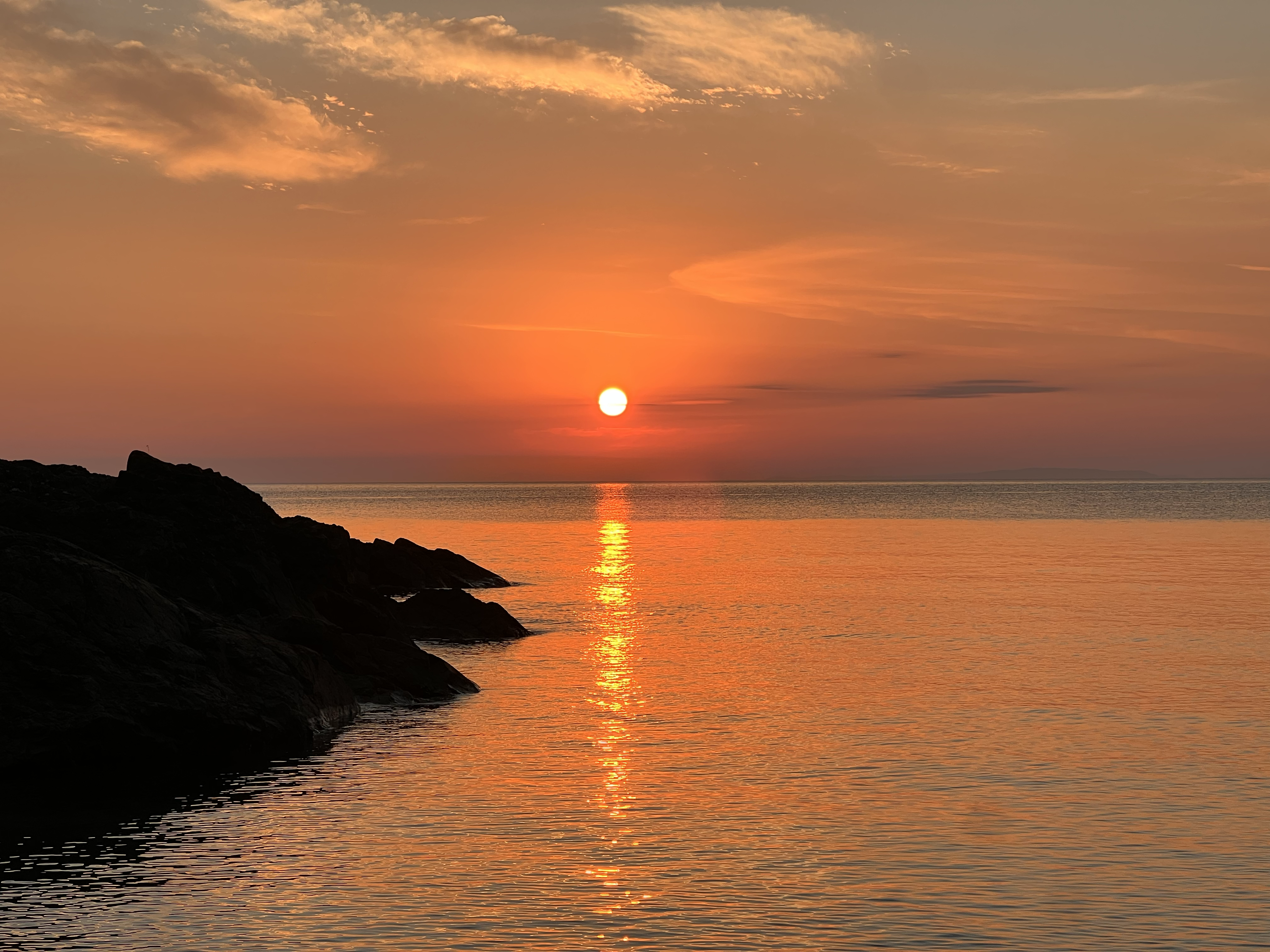 Un soleil d'un orange flamboyant plonge sous les eaux calmes du lac Supérieur, illuminant l'eau et le ciel d'une lueur orangée et dessinant la silhouette de la rive rocheuse et accidentée.