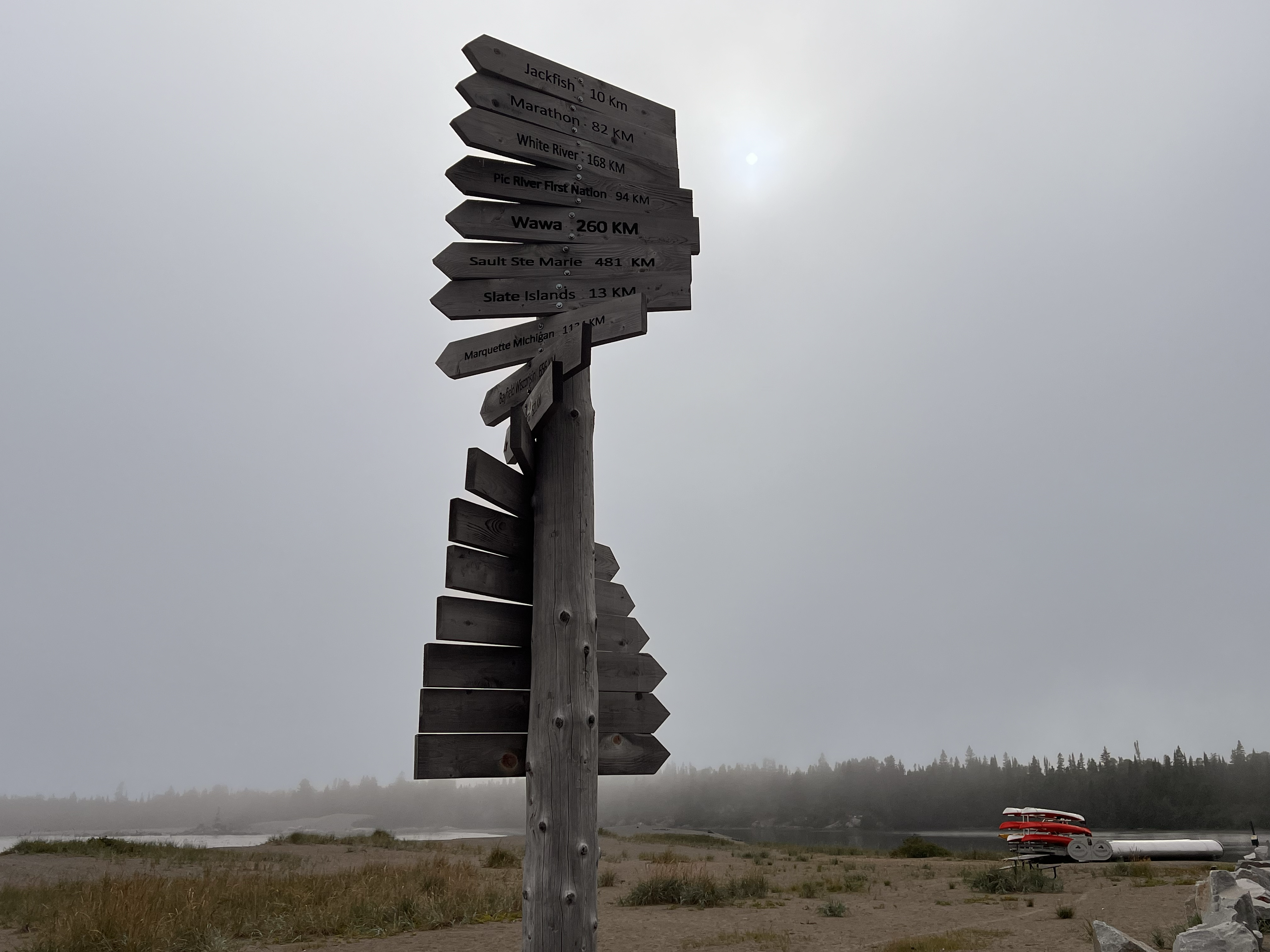 Un panneau en bois patiné par le temps, composé de plusieurs traverses, indique la direction et les distances vers les villes voisines. À l'arrière-plan, on aperçoit d'épais nuages ​​gris menaçants et une plage brumeuse où des kayaks sont chargés sur une remorque.