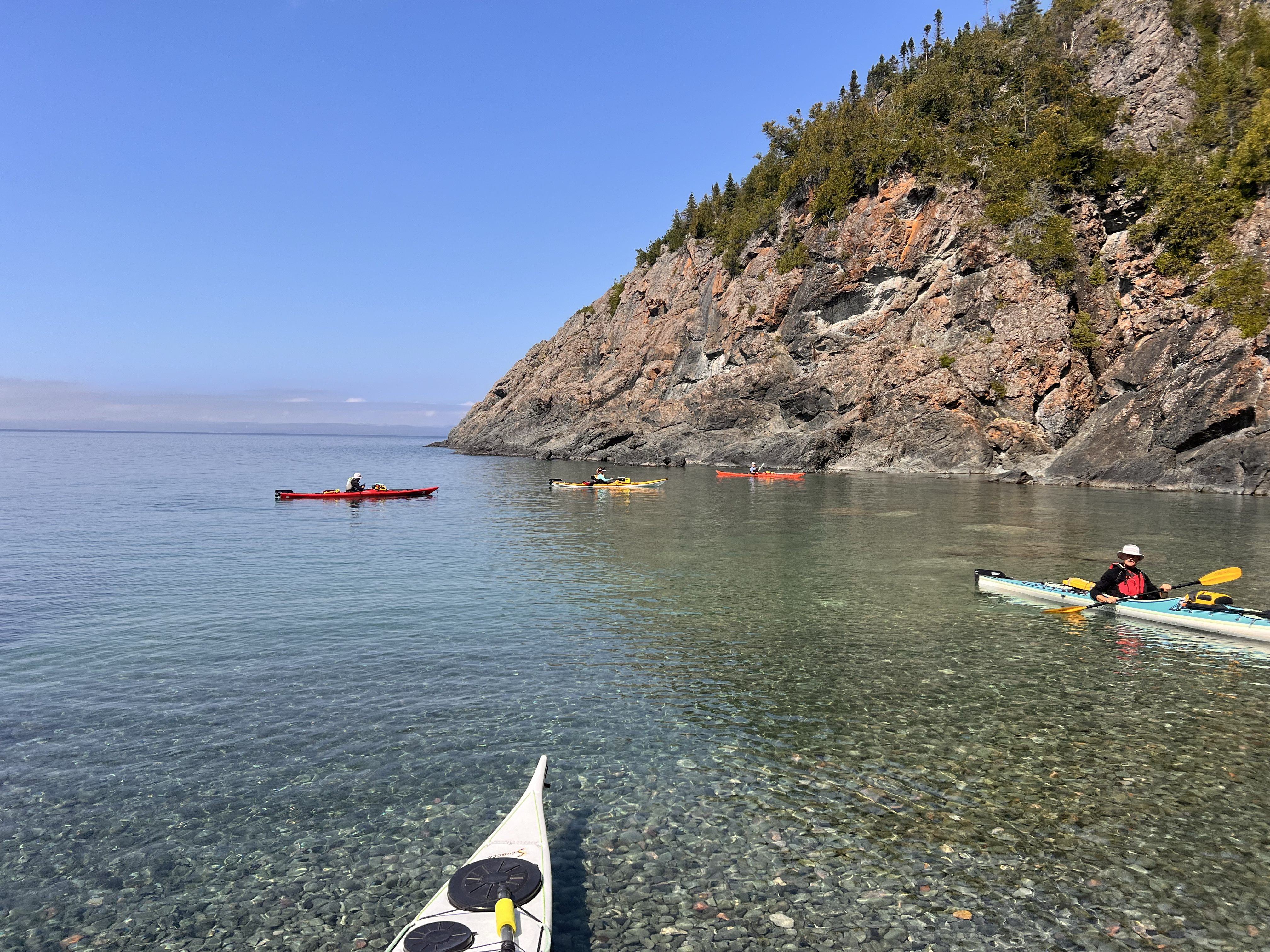 Plusieurs personnes en kayak flottent sur une baie limpide et lisse comme un miroir, à côté d'une paroi rocheuse rouge abrupte surmontée d'arbres.