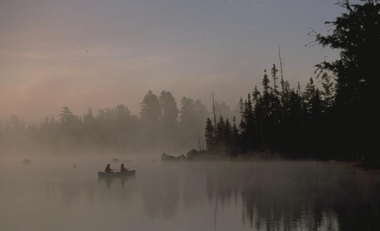 A canoe with two paddlers travelling on a lake on the misty morning. 