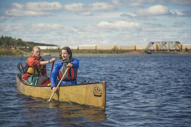 Man and woman paddling on a lake, with train in background. 