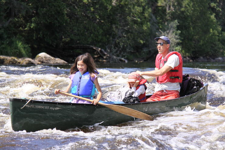 Man and two children in a green canoe paddling in gentle whitewater. 
