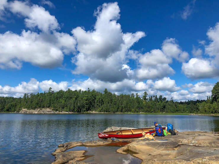 Man and dog sit beside beached canoe at edge of Lake Obabika with contents unpacked on rocks beside it